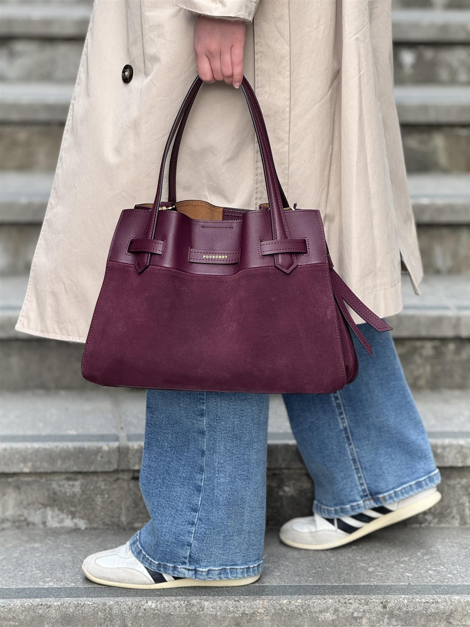 A person wearing a beige trench coat, wide-leg blue jeans, and white sneakers walks up concrete steps while holding the Blossom Burgundy large bag. A bag from Pourchet Paris.