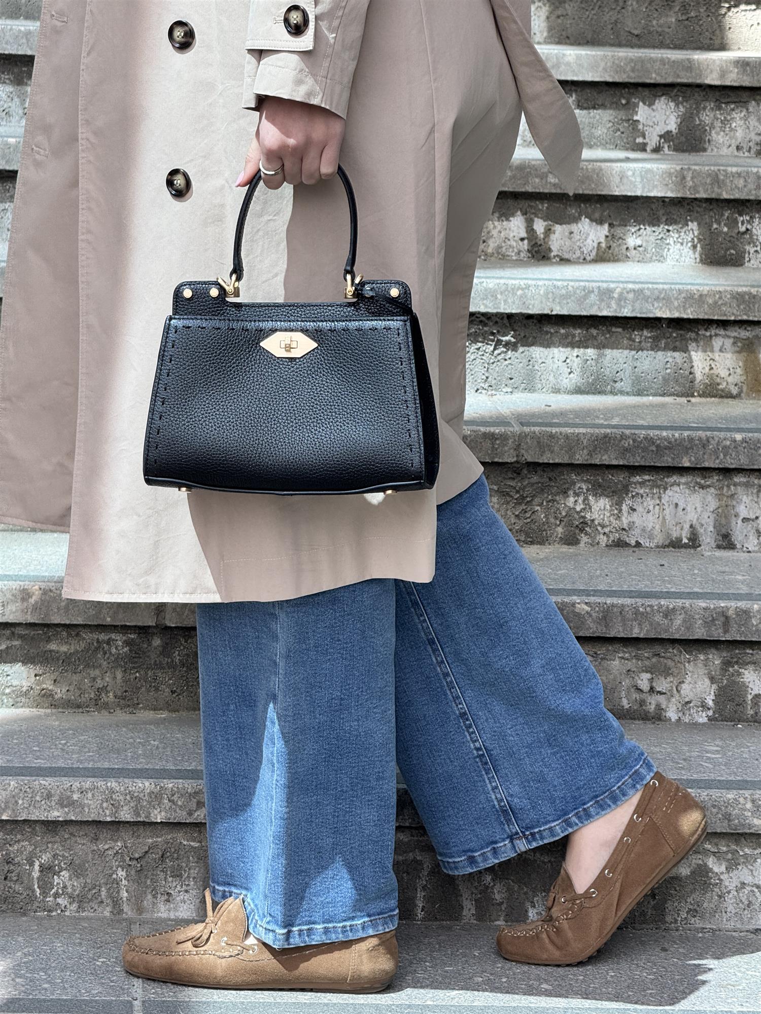A person wearing a beige trench coat, wide-leg blue jeans, and brown loafers walks up concrete steps while holding the Trianon small bag. A structured black leather handbag from Pourchet Paris.